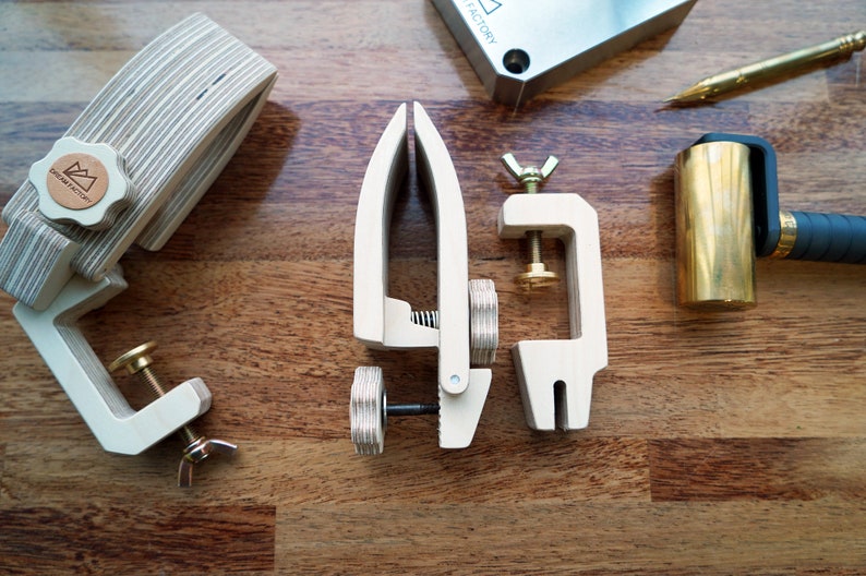 May include: Three wooden clamps with brass accents, a brass hammer with a black handle, and a metal block with the text "FACTORY" on it. The clamps are on a wooden surface.