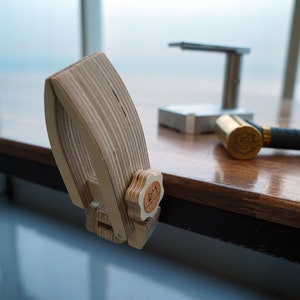 May include: A wooden clamp with a star-shaped knob, designed to hold a piece of metal in place. The clamp is on a wooden table with a brass hammer and a metal tool in the background.