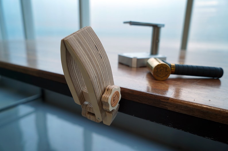 May include: A wooden clamp with a star-shaped knob, designed to hold a piece of metal in place. The clamp is on a wooden table with a brass hammer and a metal tool in the background.