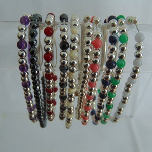 Può includere: Una collezione di 12 braccialetti con perline d'argento e pietre preziose di vari colori. I braccialetti sono esposti su un supporto trasparente.