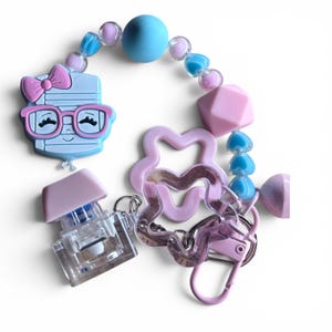 Pode incluir: Um porta-chaves com um pendente de silicone azul-claro em forma de caderno com um laço e óculos cor-de-rosa, acentuado por um interruptor de teclado mecânico transparente, contas cor-de-rosa e azuis e dois pendentes em forma de estrela ondulada.