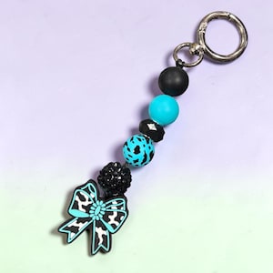 Sleutelhanger met kralen en strik in turquoise koeienprint | Tashanger van turquoise en zwart acryl | Boho sleutelring