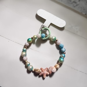 May include: A circular beaded keychain featuring a light green metal ring attached to a white display card. The beads include white and blue marbled spheres, iridescent teal and cream pearls, small gold spacers, a sea turtle, a starfish, and a shell.