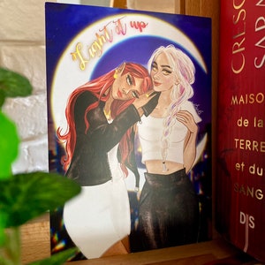 Peut inclure: Deux femmes aux cheveux longs, l'une aux cheveux roux et l'autre aux cheveux blancs, s'embrassent sous un croissant de lune. L'arrière-plan est un ciel bleu foncé avec des étoiles. Le texte "Light it up" est écrit en dégradé jaune, orange et rouge sur la lune.