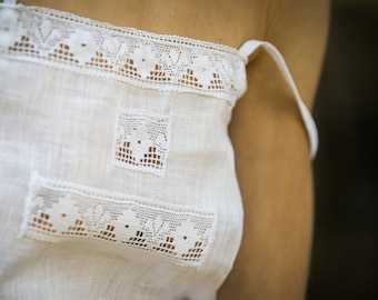 May include: Close-up of a white linen top with delicate lace trim. The top features a square lace patch and rectangular lace accents. The straps are thin and white. The fabric appears to be slightly textured.