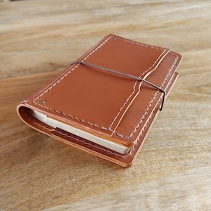 Op de afbeelding: Een bruin leren notitieboek met witte stiksels en een bruine elastische sluiting. Het notitieboek is rechthoekig en lijkt gesloten. De omslag heeft een flap en ligt op een houten oppervlak.
