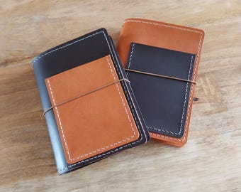 May include: Two leather notebook covers, one black and one brown, with a stitched edge and a pocket on the front. The covers are stacked on top of each other.