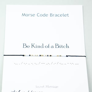 May include: A white card with black text that reads "Morse Code Bracelet" and "Be Kind of a Bitch". Below the text is a black string bracelet with white and gold beads. The bracelet is a Morse code bracelet with the message "Be Kind of a Bitch" spelled out in Morse code. The card also includes the text "Secret Message" and "Wishanddesign" with a website address.