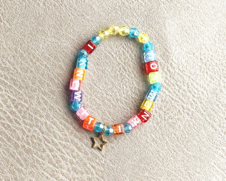 May include: A circular bracelet made of multicolored translucent faceted beads and square letter beads spelling out I KNEW IT. The bracelet features a small, gold-toned metal star charm dangling from the bottom, interspersed with blue, yellow, pink, and red beads.