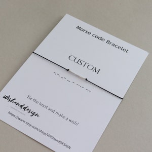 以下が含まれることがあります： 黒の紐のブレスレットに、銀色のビーズがモース信号でメッセージを綴っています。ブレスレットは、白いカードの上に置かれており、カードには「モールス信号ブレスレット CUSTOM 結び目を結び、願いを込めて！ Wishanddesign 手作りのブレスレット https://www.etsy.com/shop/WISHandDESIGN」と書いてあります。