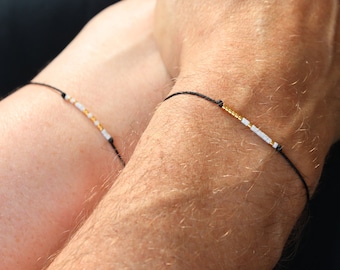 May include: Two black string bracelets, each adorned with a series of small, colorful beads. One bracelet features gold and white beads, while the other showcases a similar pattern. The bracelets are displayed on wrists.