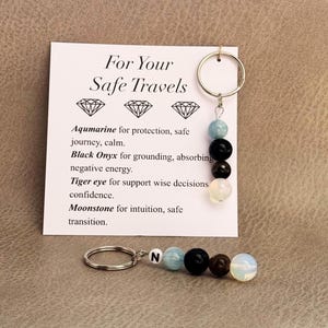 May include: Two keychains featuring a silver metal ring and a strand of four round beads in light blue, black, dark brown, and iridescent white. One keychain includes a white bead with the letter N. A white card displays the text For Your Safe Travels.