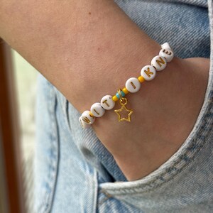 May include: A beaded bracelet featuring white circular beads with gold letters spelling WITI OKNE, interspersed with small yellow and turquoise seed beads. A gold-toned metal star charm hangs from the center of the bracelet, which is worn on a wrist.