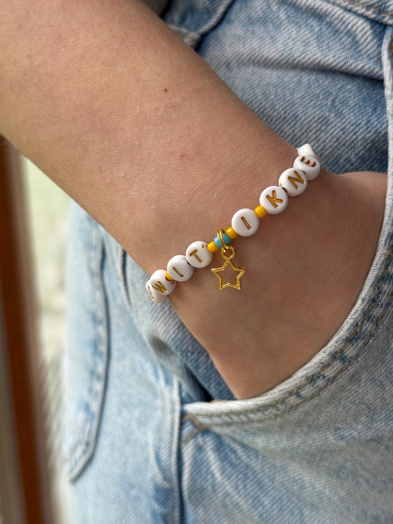 May include: A beaded bracelet featuring white circular beads with gold letters spelling WITI OKNE, interspersed with small yellow and turquoise seed beads. A gold-toned metal star charm hangs from the center of the bracelet, which is worn on a wrist.