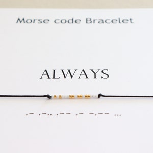 Pode incluir: Um bracelete de corda preta com contas brancas e douradas que soletram a palavra "SEMPRE" em código Morse. O bracelete é exibido em um cartão branco com as palavras "Bracelete de código Morse" e "SEMPRE" impressas nele.