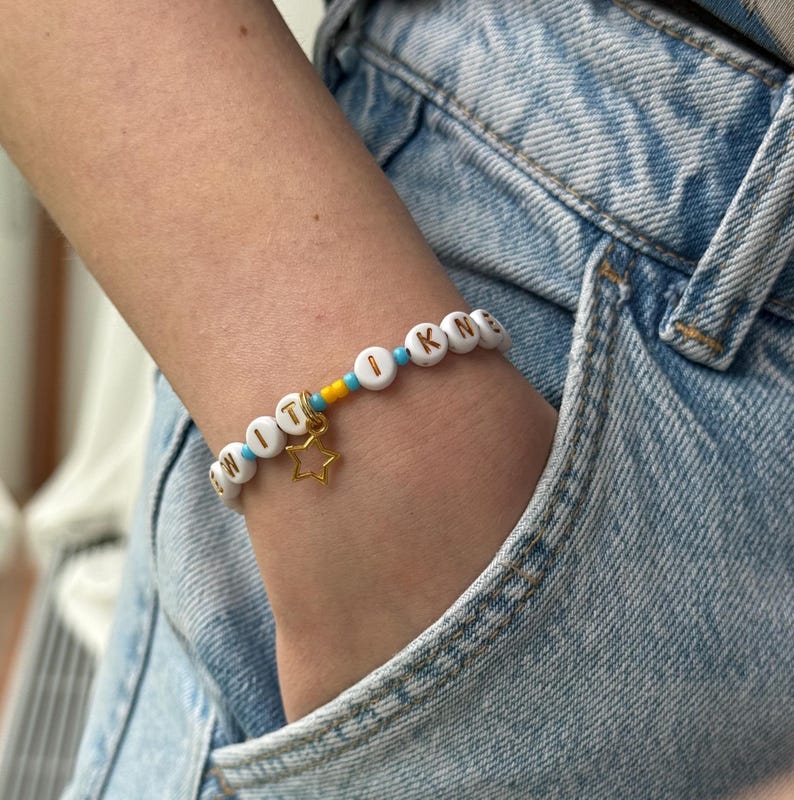 May include: A beaded bracelet featuring white round beads with black letters spelling out a word, interspersed with small turquoise and yellow spacer beads. A gold-toned metal star charm hangs from the center of the bracelet string.
