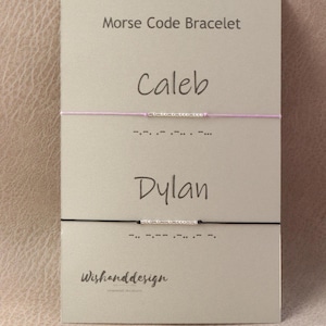 May include: Two thin cord bracelets on a beige display card labelled Morse Code Bracelet. One bracelet is light purple with silver beads spelling Caleb, the other is black with silver beads spelling Dylan. Both include printed Morse code dots and dashes.