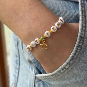May include: A beaded bracelet featuring white circular beads with gold letters spelling WITOKNE, interspersed with small yellow and turquoise seed beads. A gold-toned open star charm hangs from the center of the bracelet, which is worn on a wrist.