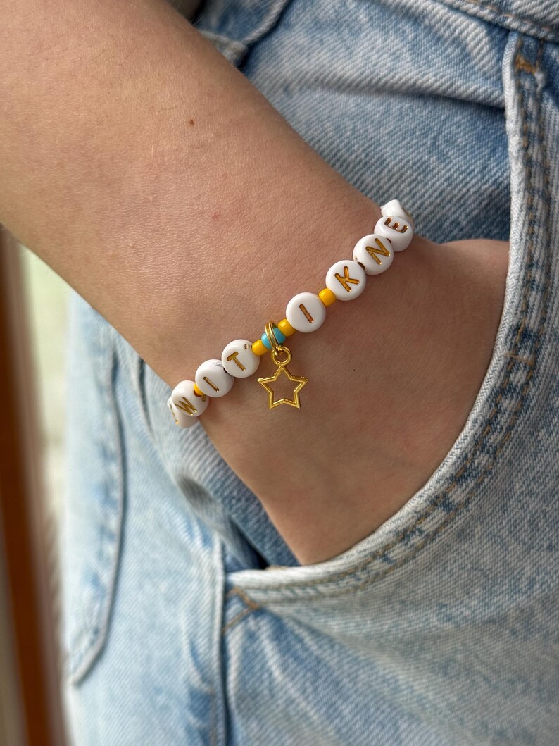 May include: A beaded bracelet featuring white circular beads with gold letters spelling WITOKNE, interspersed with small yellow and turquoise seed beads. A gold-toned open star charm hangs from the center of the bracelet, which is worn on a wrist.