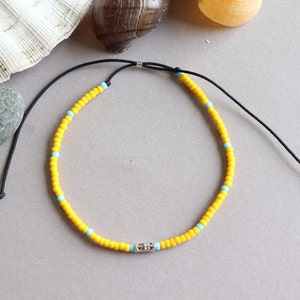 以下が含まれることがあります： 銀色の留め具が付いた黄色と青のビーズのアンクレット。アンクレットは小さな丸いビーズで作られており、黒い紐で結ばれています。