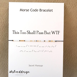 Puede incluir: Una pulsera de cordón negro fino con pequeñas cuentas blancas, negras y doradas dispuestas en un patrón de código Morse. La pulsera se presenta sobre una tarjeta blanca con el texto Morse Code Bracelet, This Too Shall Pass But WTF y Secret Message.