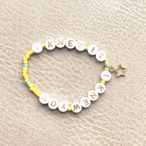 May include: A beaded bracelet featuring white circular beads with gold lettering that spells out I KNEW IT I KNEW YOU. The design includes smaller yellow and light blue seed beads interspersed throughout the band and a small gold star charm.