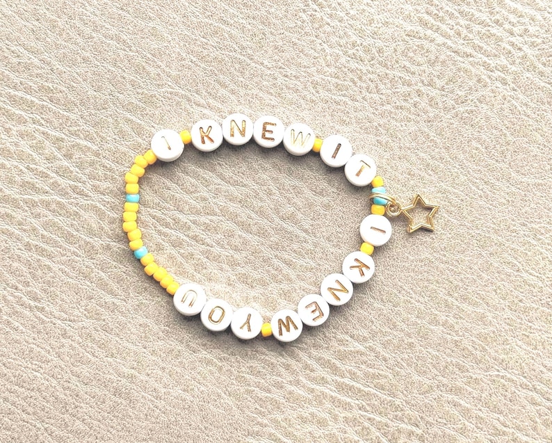 May include: A beaded bracelet featuring white circular beads with gold lettering that spells out I KNEW IT I KNEW YOU. The design includes smaller yellow and light blue seed beads interspersed throughout the band and a small gold star charm.