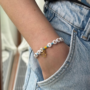 May include: A beaded bracelet featuring white circular beads with black letters spelling out text, interspersed with small bright blue and yellow beads. A gold-toned metal star charm hangs from the center of the bracelet, which is worn on a wrist.