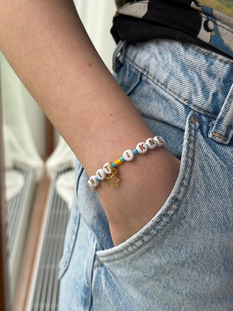 May include: A beaded bracelet featuring white circular beads with black letters spelling out text, interspersed with small bright blue and yellow beads. A gold-toned metal star charm hangs from the center of the bracelet, which is worn on a wrist.