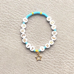 May include: A circular beaded bracelet featuring light blue and yellow seed beads interspersed with white round beads. The white beads display the gold-colored text I KNEW IT I KNEW IT. A small, open-frame gold-colored star charm hangs from the bottom.