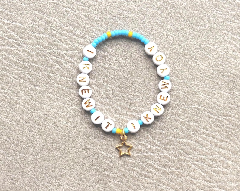 May include: A circular beaded bracelet featuring light blue and yellow seed beads interspersed with white round beads. The white beads display the gold-colored text I KNEW IT I KNEW IT. A small, open-frame gold-colored star charm hangs from the bottom.