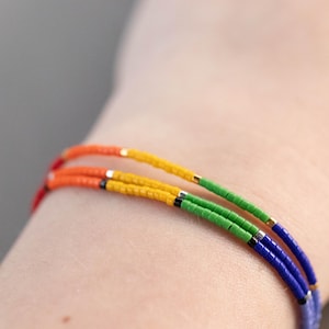 Peut inclure: Un bracelet multi-rangs coloré. Le bracelet est composé de rangs de perles rouges, oranges, jaunes, vertes et bleues. Le bracelet est porté sur un poignet.