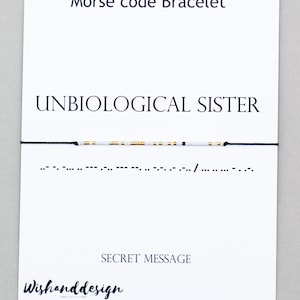 Könnte beinhalten: Weißes und goldenes Perlenarmband mit schwarzem Band. Das Armband buchstabiert "UNBIOLOGICAL SISTER" in Morsecode. Der Text "SECRET MESSAGE" ist unter dem Armband gedruckt.