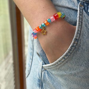 May include: A colorful beaded bracelet featuring square translucent beads with white letters, interspersed with small blue faceted beads. The bracelet includes a gold-toned star charm hanging from the center, creating a vibrant accessory.