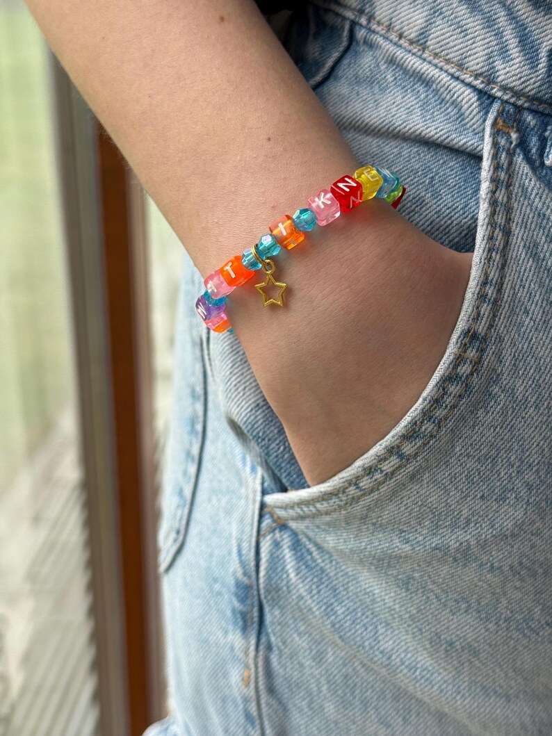 May include: A colorful beaded bracelet featuring square translucent beads with white letters, interspersed with small blue faceted beads. The bracelet includes a gold-toned star charm hanging from the center, creating a vibrant accessory.