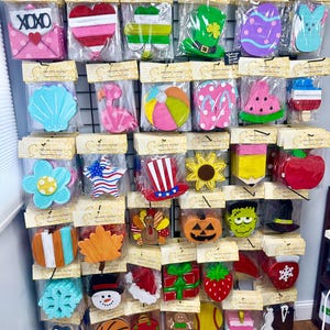 May include: A collection of assorted seasonal wooden cutout decorations packaged in clear plastic bags with Creative Sisters headers. The items feature various shapes including hearts, flowers, holiday symbols, food, and sports themes in vibrant colors.