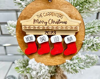 May include: A wooden Christmas ornament with a round shape. It features the text "The Carpenters Merry Christmas 2023" and four red stockings labeled "Mom," "Dad," "Leo," and "Ava." The ornament hangs from a red and white string.