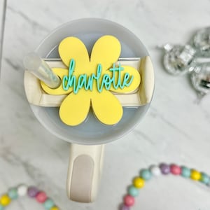 May include: A yellow flower-shaped lid with the name "charlotte" written in teal on it. The lid is on top of a white cup with a handle.