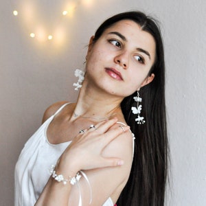 May include: A woman with long dark hair wearing a white tank top and white flower earrings. She is wearing a white bracelet with a silver charm on her wrist. She is looking to the side and smiling.