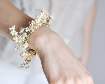May include: A wrist adorned with a delicate bracelet made of dried white flowers and greenery. The bracelet is tied with a white ribbon. The person is also wearing several silver rings on their fingers.