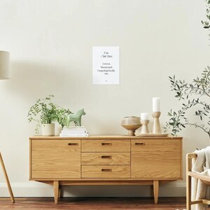 Könnte beinhalten: Ein Sideboard aus hellem Holz mit drei Schubladen und zwei Türen, das Dekorationsgegenstände enthält. Ein gerahmter Druck mit Text hängt an der Wand. Eine Stehlampe und ein Stuhl befinden sich ebenfalls im Raum. Eine kleine grüne Pferdfigur sitzt auf Büchern.