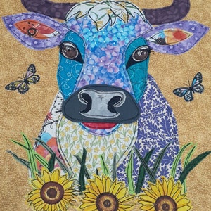 Pode incluir: Uma vaca de patchwork azul e roxo com nariz preto e lábios vermelhos, cercada por girassóis amarelos e grama verde. Há também três borboletas na imagem, duas azuis e uma preta.