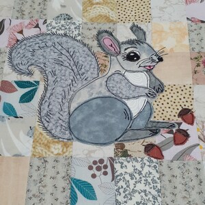 May include: An embroidered grey and white squirrel with a bushy tail, sitting on a patchwork quilt with various floral and geometric patterns. The quilt is made of different fabrics in shades of brown, beige, pink, and white.