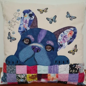 Peut inclure: Un oreiller blanc avec un bouledogue français brodé bleu et violet qui dépasse d'un patchwork de carrés de tissu colorés. L'oreiller a le mot "Hope" brodé en fil bleu dans le coin supérieur gauche.