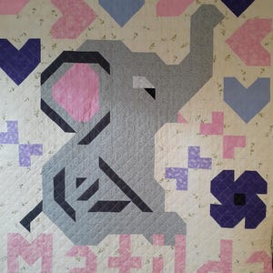 May include: A quilted baby blanket featuring a gray elephant with a pink ear, surrounded by pink and purple shapes. The name "Matilda" is spelled out in pink letters at the bottom. The background is a cream color with a floral pattern.