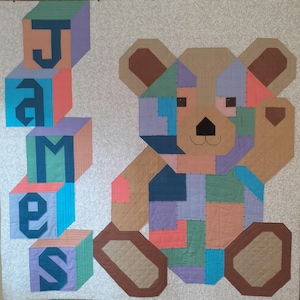Op de afbeelding: Een handgemaakte quilt met een geometrisch teddybeerontwerp met een patchwork van verschillende kleuren. De naam "JAMES" is in grote, kleurrijke blokken gespeld. De quilt heeft een lichte achtergrond.