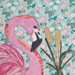 Puede incluir: Un flamenco rosa con pico y ojo negro y blanco sobre un fondo floral. El flamenco está rodeado de juncos y hojas verdes. El fondo es una tela azul claro con flores rosas y blancas.