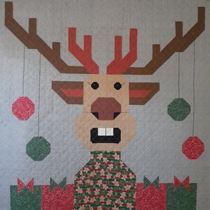 May include: A Christmas-themed quilt featuring a reindeer design. The reindeer has brown antlers, a tan face, and a green and red patterned sweater. Red and green ornaments hang from the antlers. The quilt has a gray background and a quilted texture.