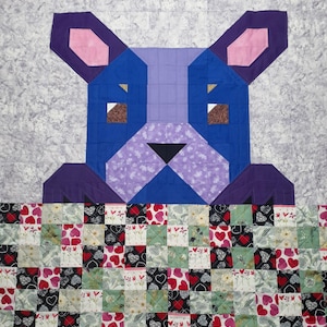 Może przedstawiać: Ręcznie robiona kołdra z geometrycznym wzorem niebieskiej psiej twarzy z fioletowymi uszami i czarnym nosem. Głowa psa znajduje się nad patchworkową obwódką z wzorami serc i kwiatów. Kołdra wykonana jest z kwadratów materiału.