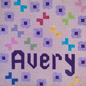 May include: A lavender quilt with a repeating pattern of colorful crosses and floral shapes. The name "Avery" is prominently displayed in a dark purple, geometric font, adding a personalized touch to the design.